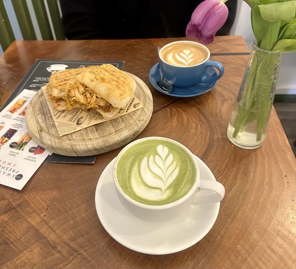 A café table set with a toasted sandwich on a board, a cappuccino, a matcha latte and tulips in a vase
