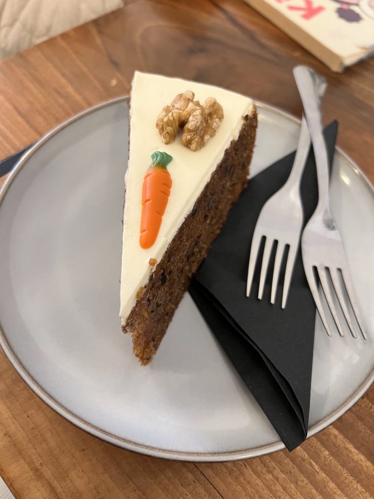 A slice of carrot cake with a marzipan carrot, plated with cutlery and a black napkin